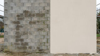 A wall split vertically: left, rough grey cinder blocks with moisture/algae; right, smooth light beige plaster, showing material contrast.