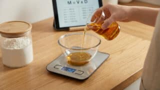 A hand pours golden liquid from a measuring cup into a bowl on a digital scale (150g) on a wooden counter. Flour jar and tablet with M=VxD in background.