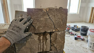 Close-up of a gloved hand holding a trowel next to a cracked stone veneer wall with visible water stains and construction tools in the background.