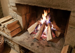 Vibrant fig wood logs burn in a rustic stone fireplace, casting a warm glow. A stack of pale, fine-grained fig wood awaits nearby.