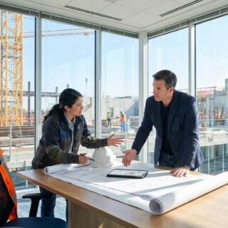 Une femme et un homme discutent de plans sur une table de bureau, avec un chantier en arrière-plan.