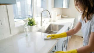 Une femme portant des gants jaunes nettoie un plan de travail blanc dans une cuisine lumineuse.
