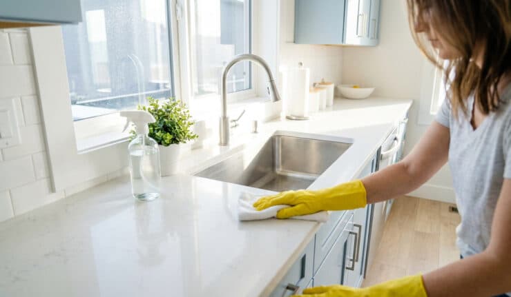 Une femme portant des gants jaunes nettoie un plan de travail blanc dans une cuisine lumineuse.